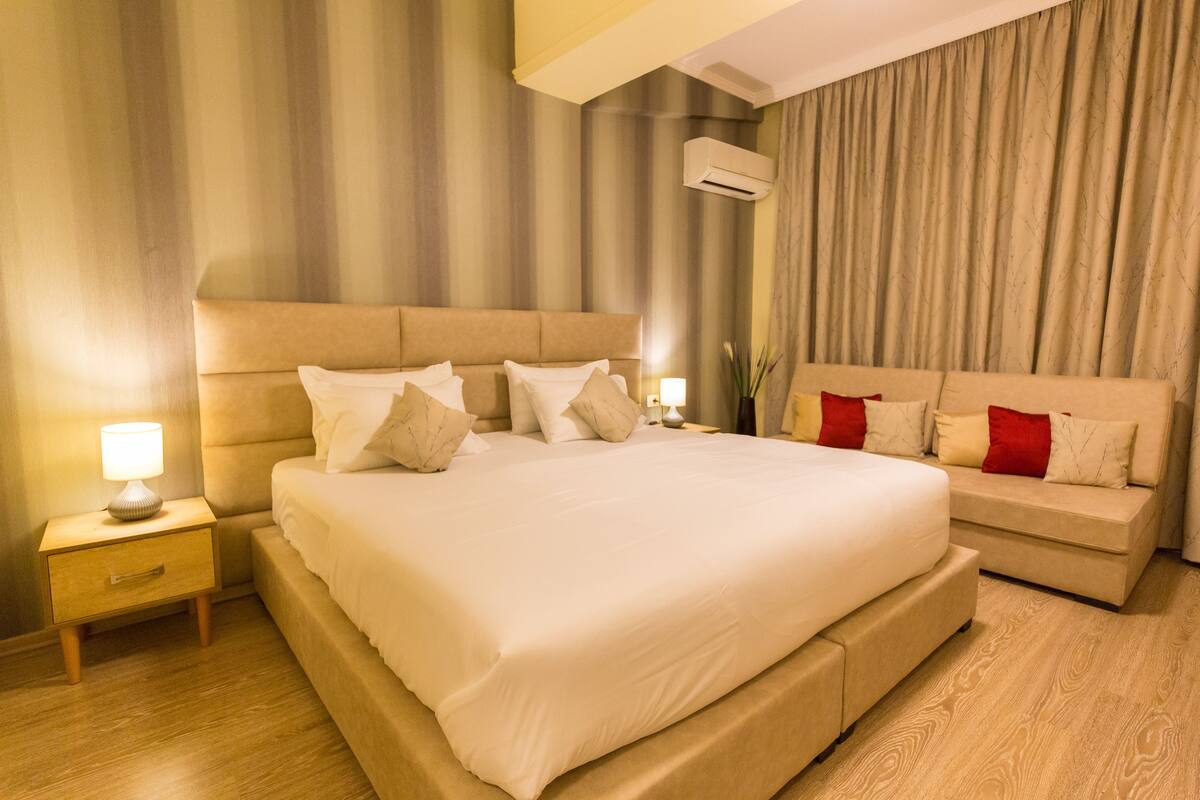 A large bed with a headboard, a bedside table with a lamp, and a sofa with cushions.