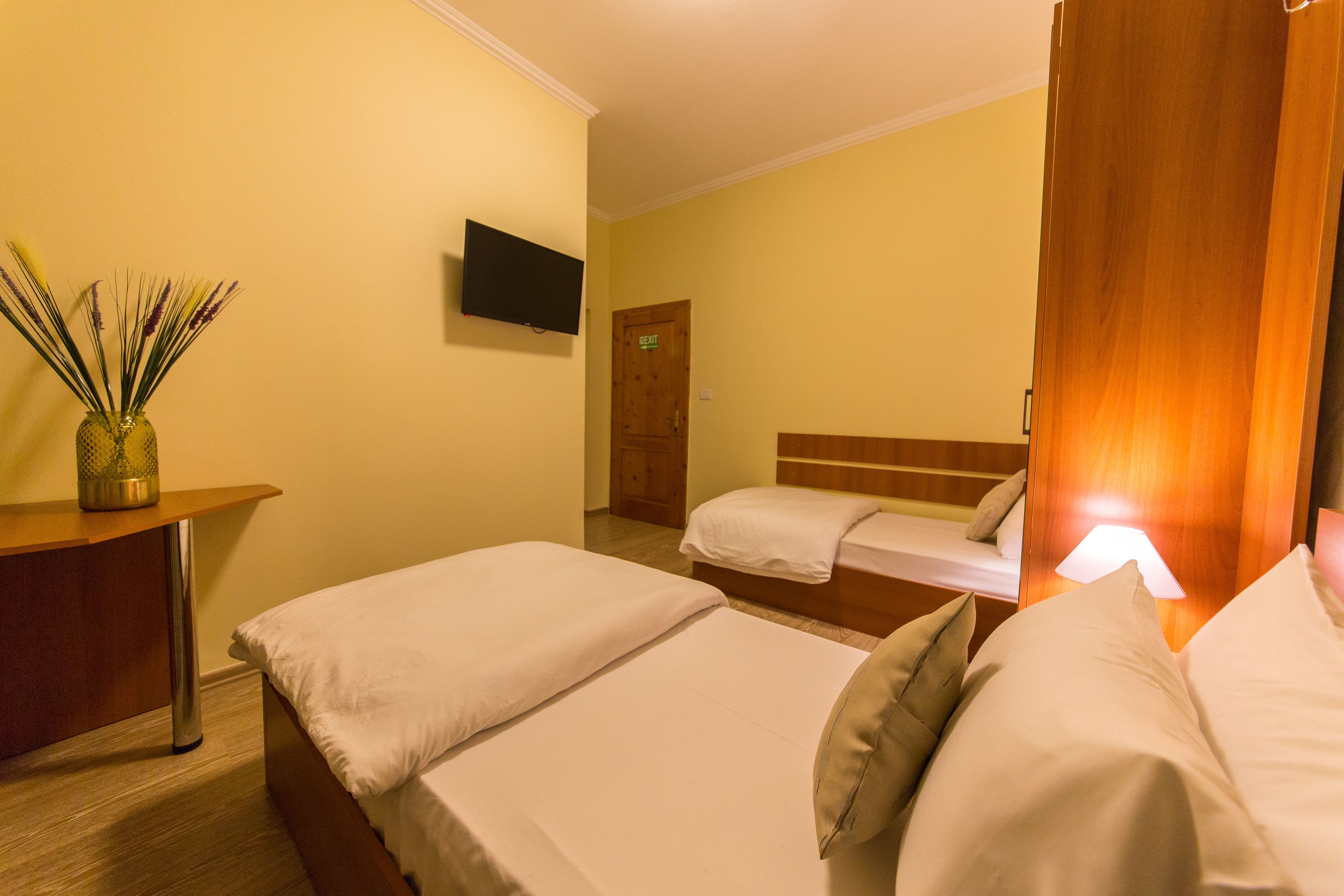 A hotel room with two beds, a TV mounted on the wall, and a wooden desk with a vase of dried plants.