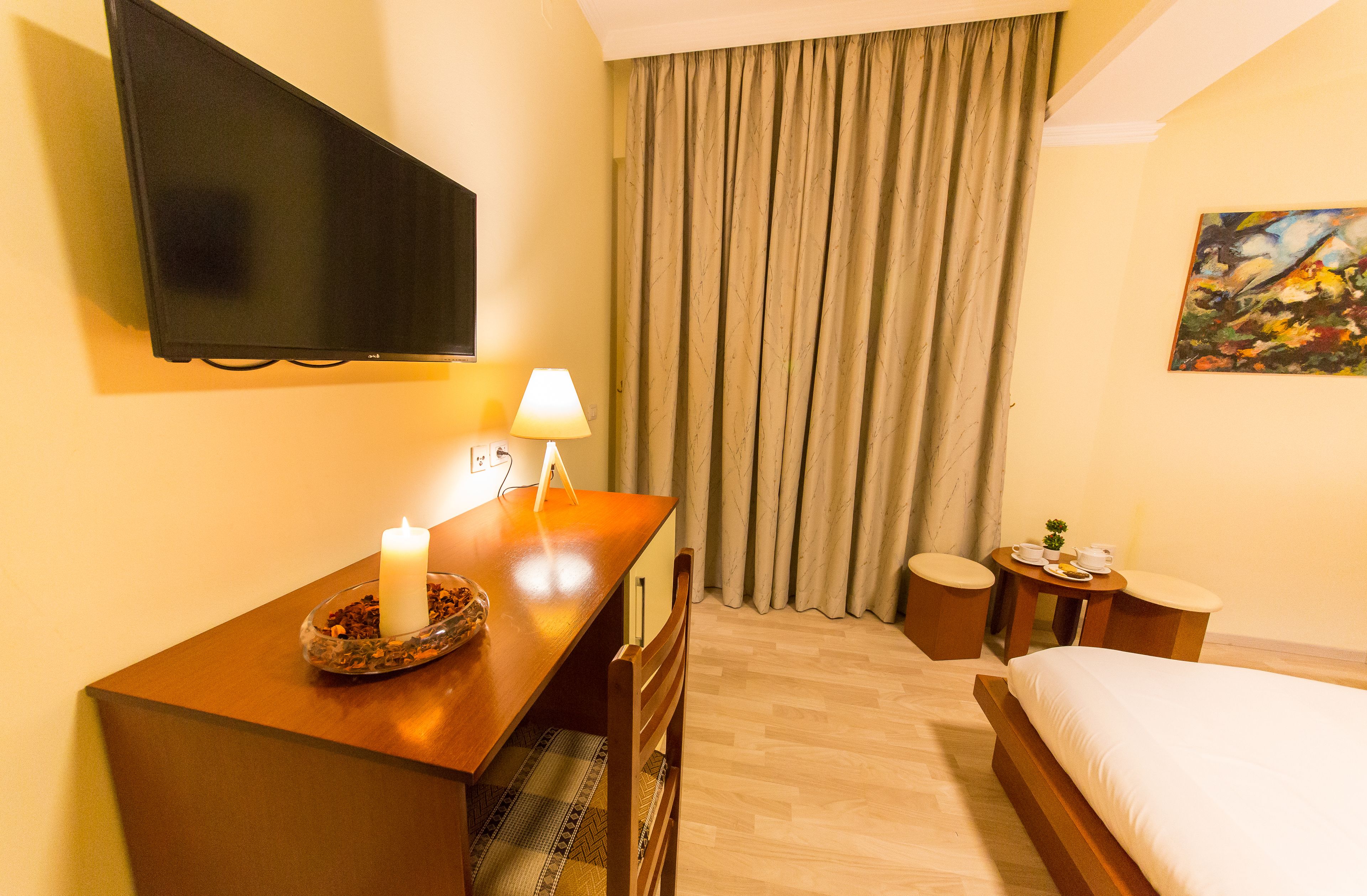 A hotel room with a bed, a desk with a lamp, a TV mounted on the wall, a painting on the wall, and a window with curtains.