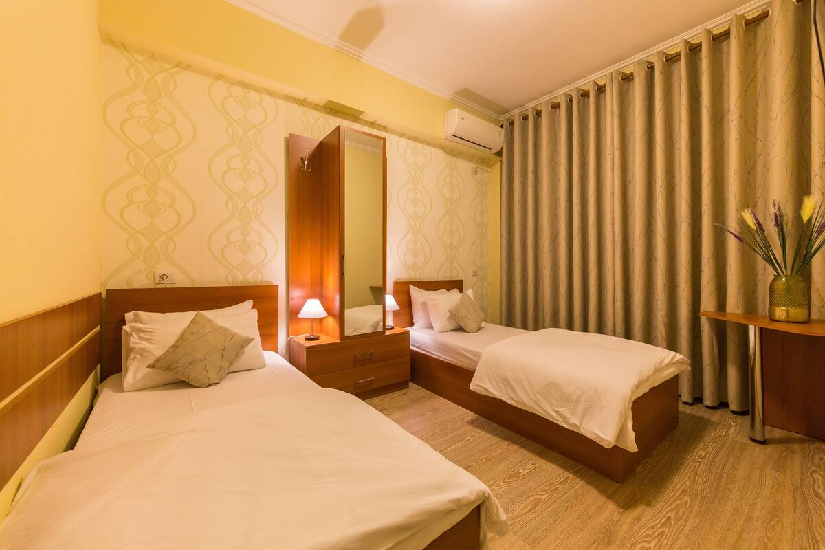 A hotel room with two beds, a wooden headboard, a nightstand with a lamp, a mirror, and a window with curtains.