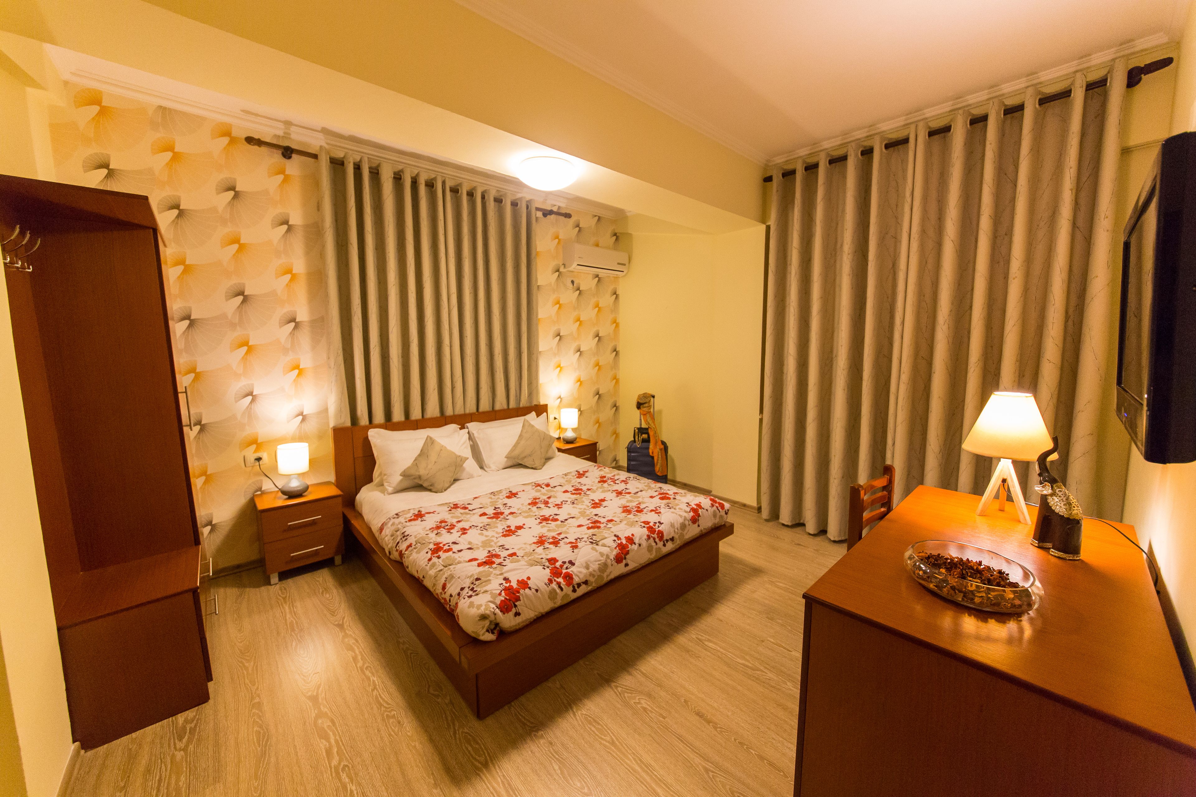 Une chambre d’hôtel avec un lit en bois, une table de chevet avec une lampe, une commode et une fenêtre avec des rideaux.