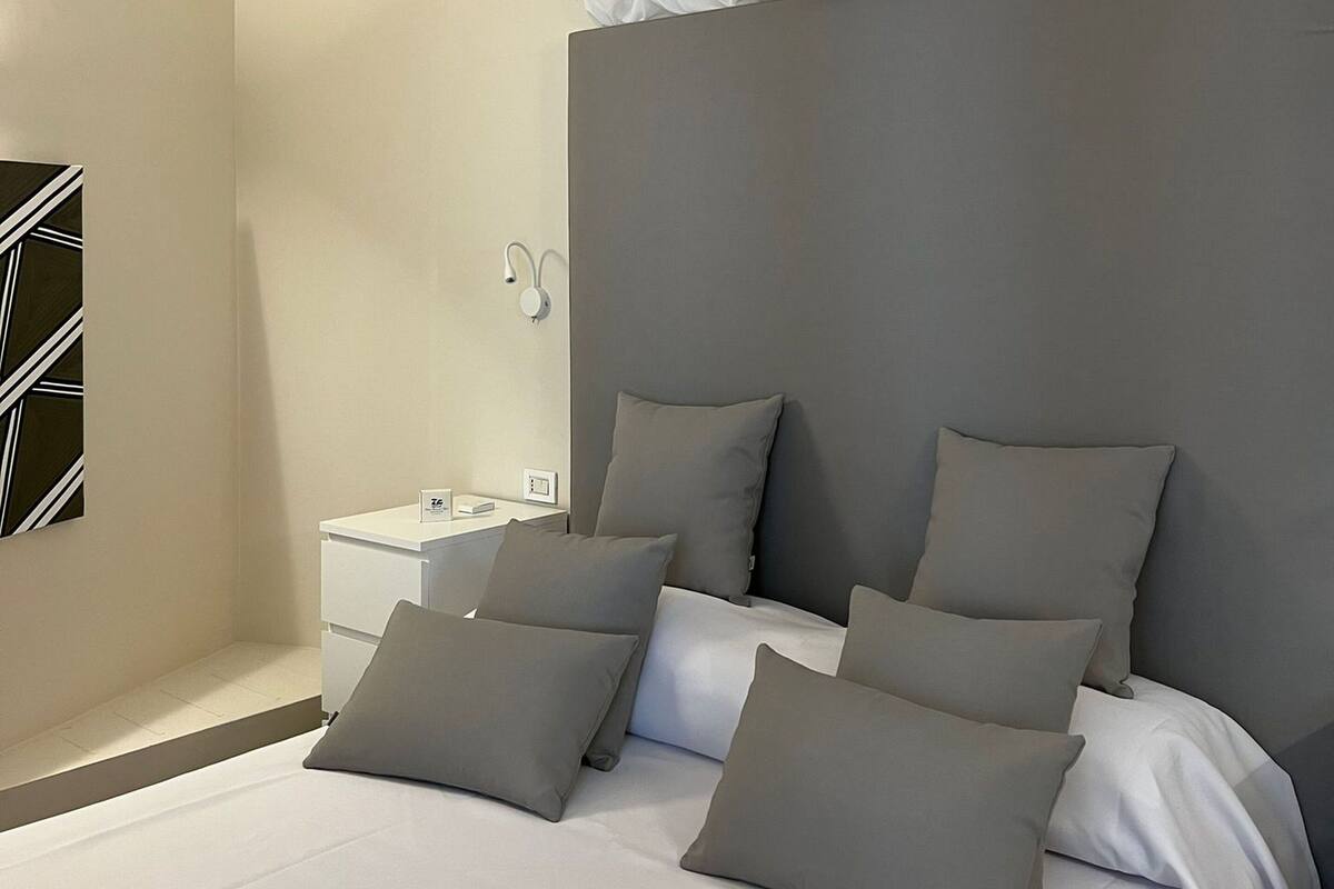 Ein modernes Schlafzimmer mit einem großen Bett, grauen Kissen, einem Nachttisch und einer Wandleuchte.