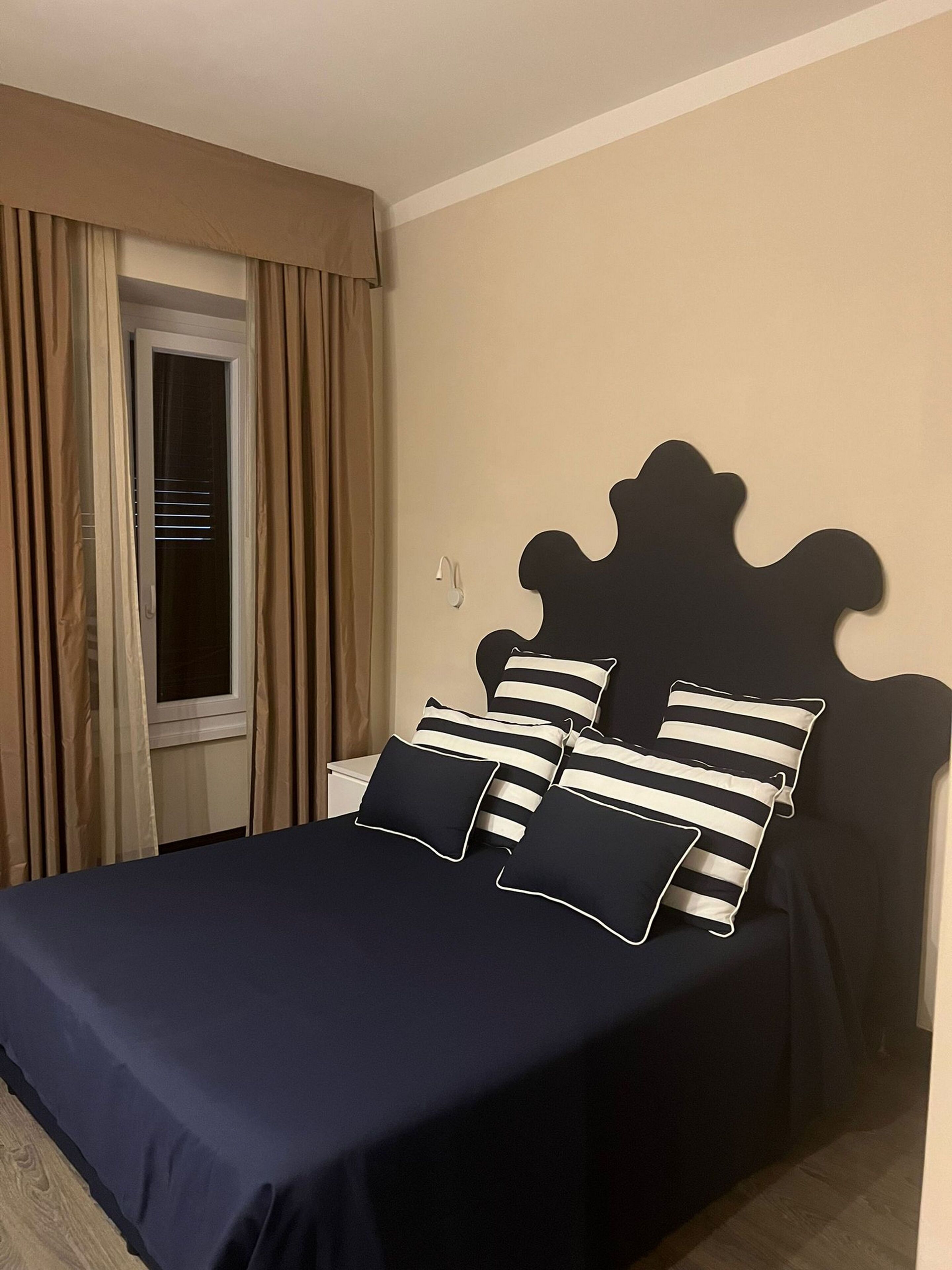 A bed with a dark blue bedspread and striped pillows, a window with curtains, and a wall-mounted light fixture.