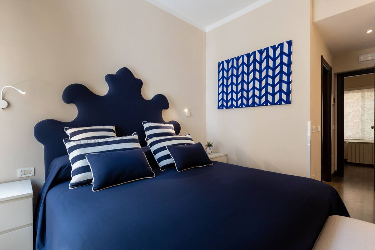 Ein Schlafzimmer mit einem dunkelblauen Bett, gestreiften Kissen, einer Wandleuchte und einem Wandbild mit geometrischem Muster.