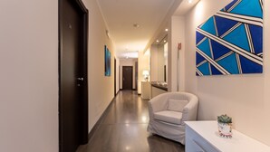 A hallway with a dark wood floor, a white armchair, a white console table with a potted plant, and a blue abstract painting on the wall. - Relais Piazza Del Popolo - Aminta Collection Luxury Rooms (Rome)