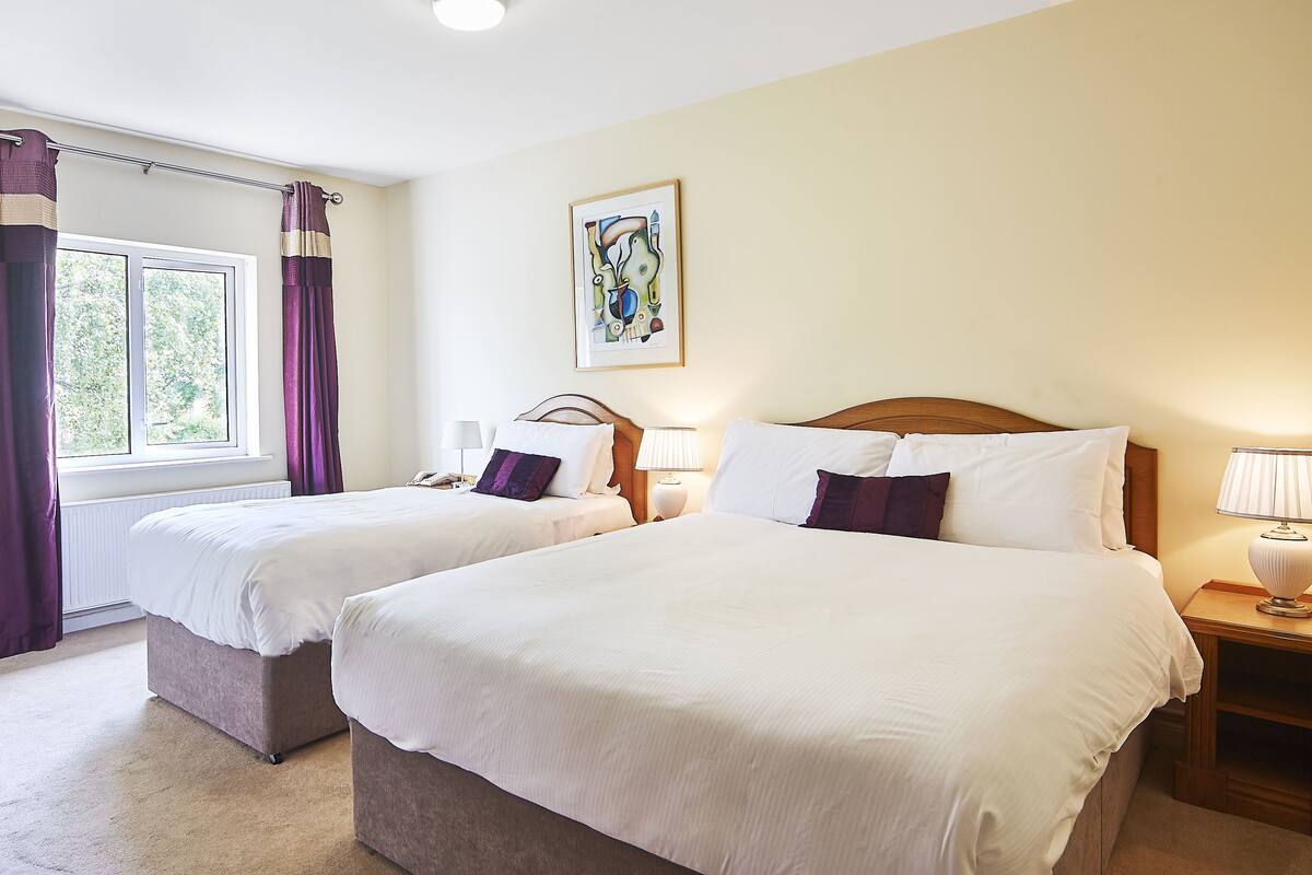 A hotel room with two beds, a window with curtains, a painting on the wall, and a bedside table with a lamp.