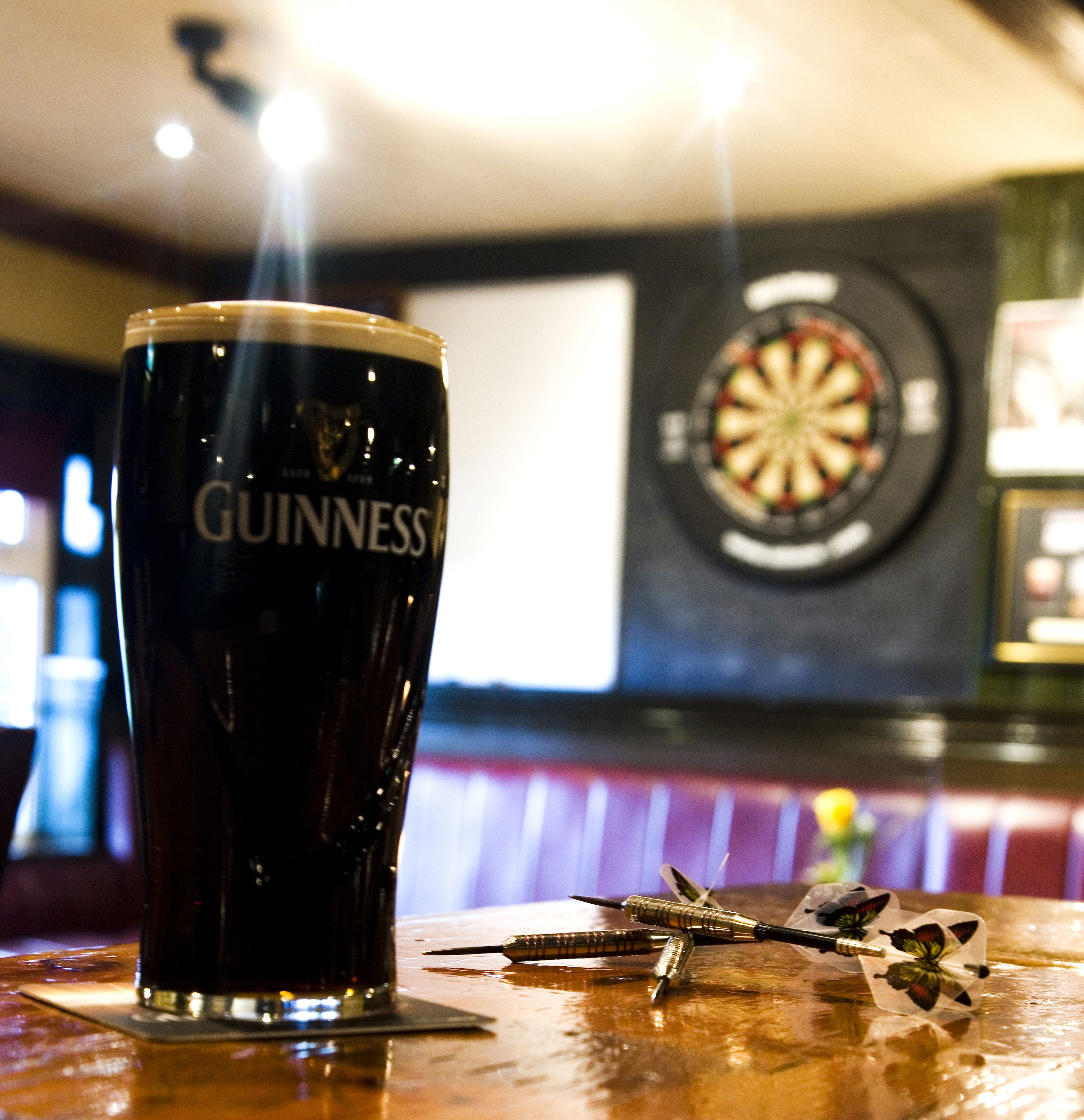 A Guinness glass on a wooden table with darts and a dartboard in the background.