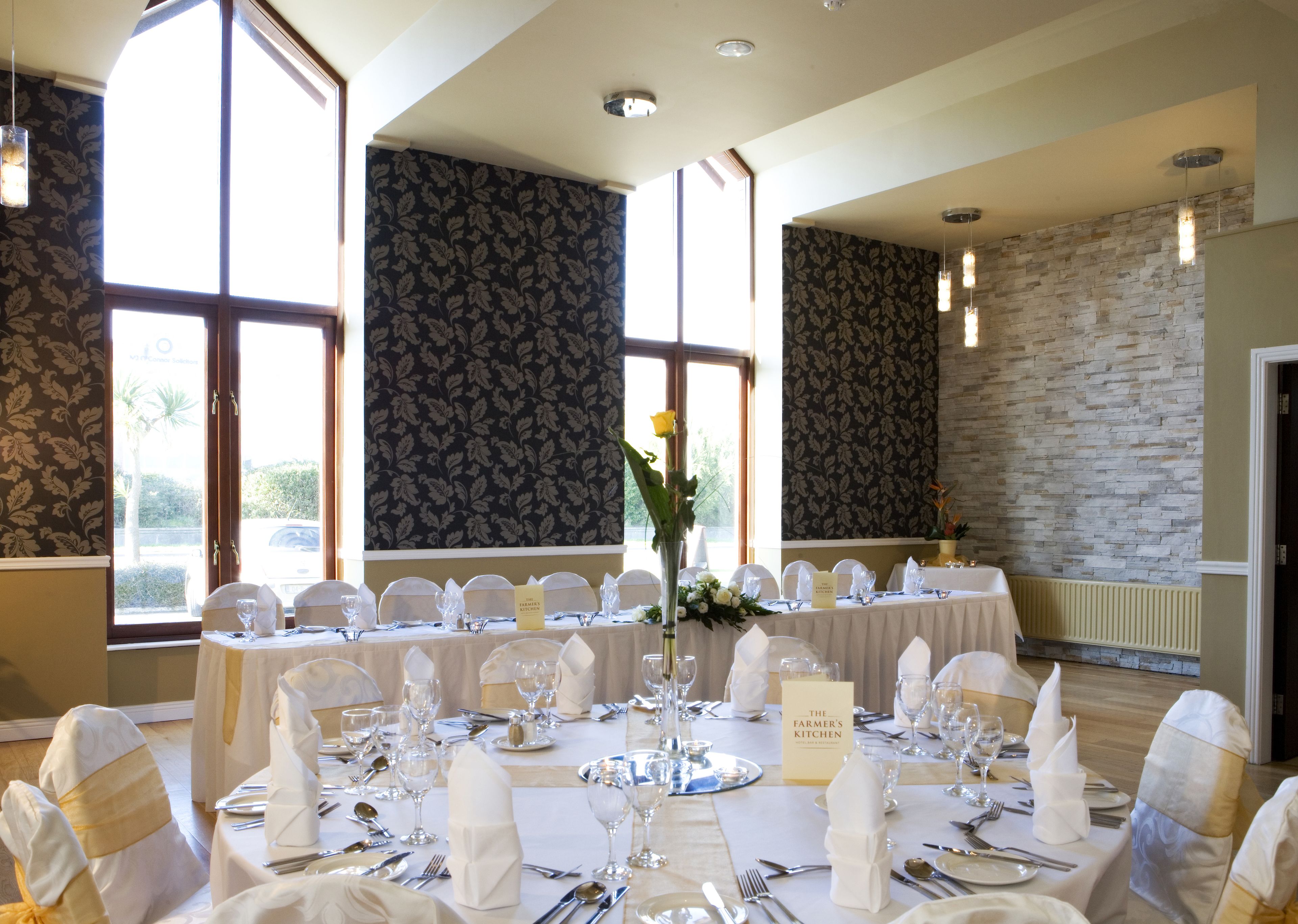 A banquet hall with tables set for a formal event, featuring a stone accent wall and large windows.