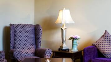 A room with a patterned armchair, a sofa, a table, a lamp, and a vase with flowers.