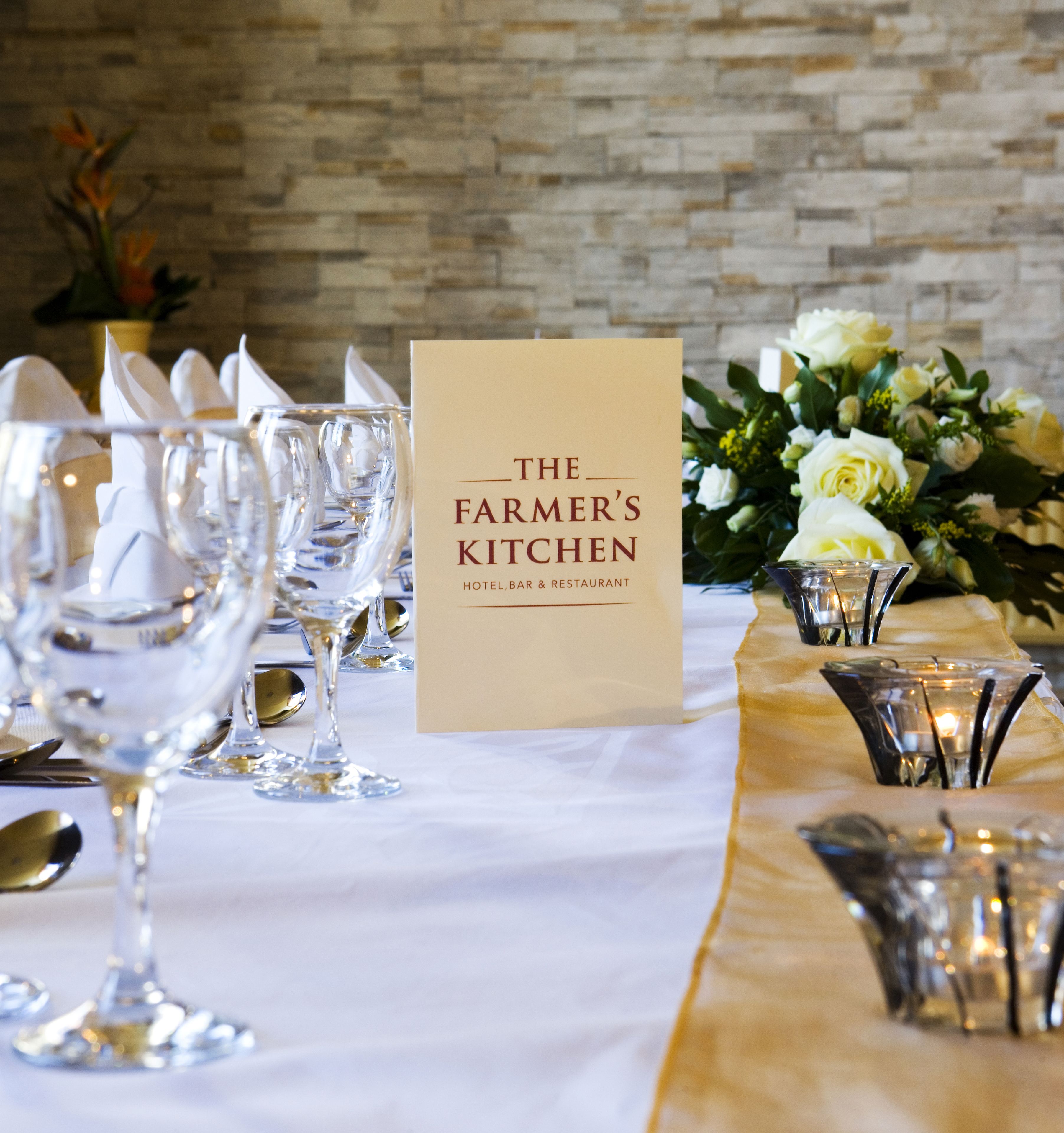 A table set with wine glasses, a menu titled "The Farmer's Kitchen," and a vase with white flowers.