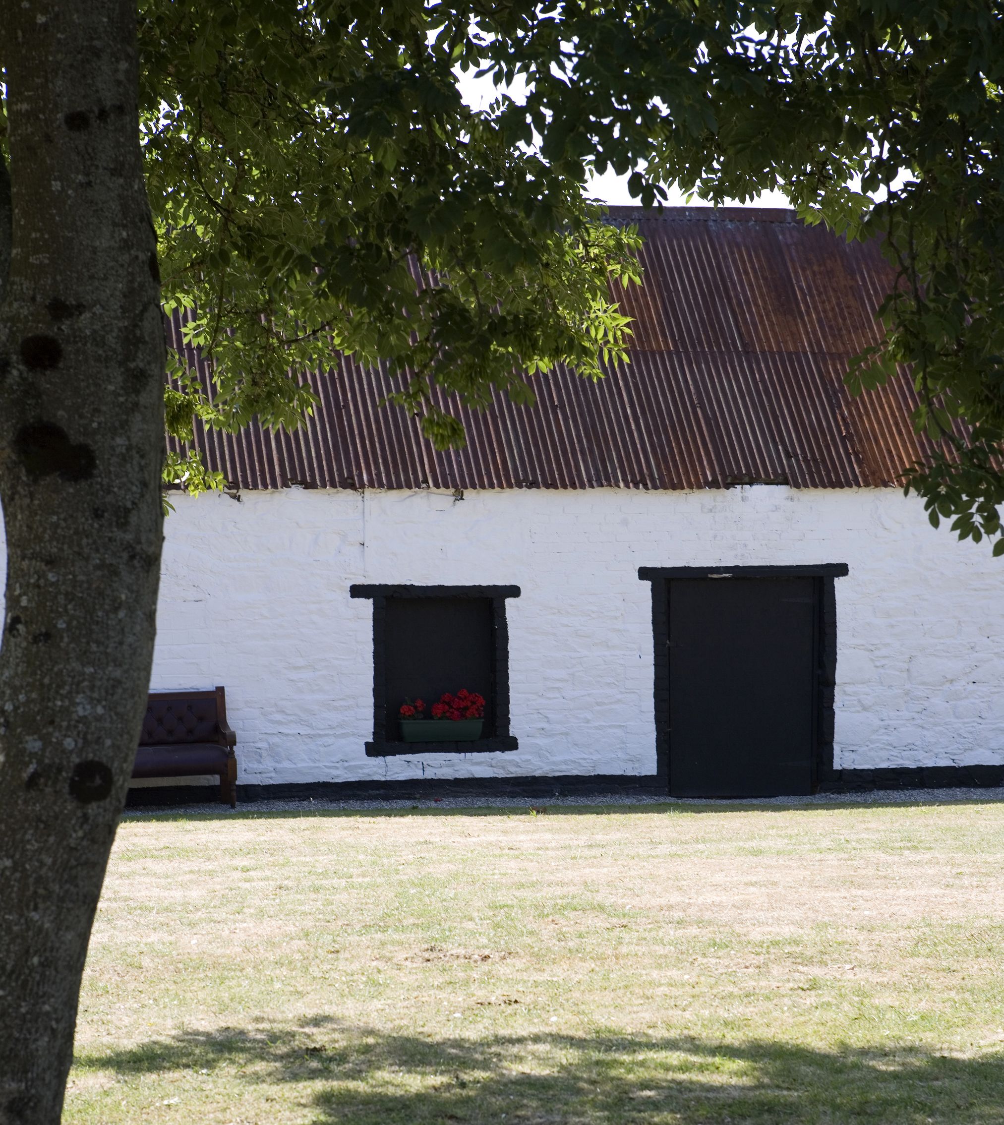 Un edificio blanco con techo de lámina corrugada, dos puertas negras y una ventana con flores rojas.