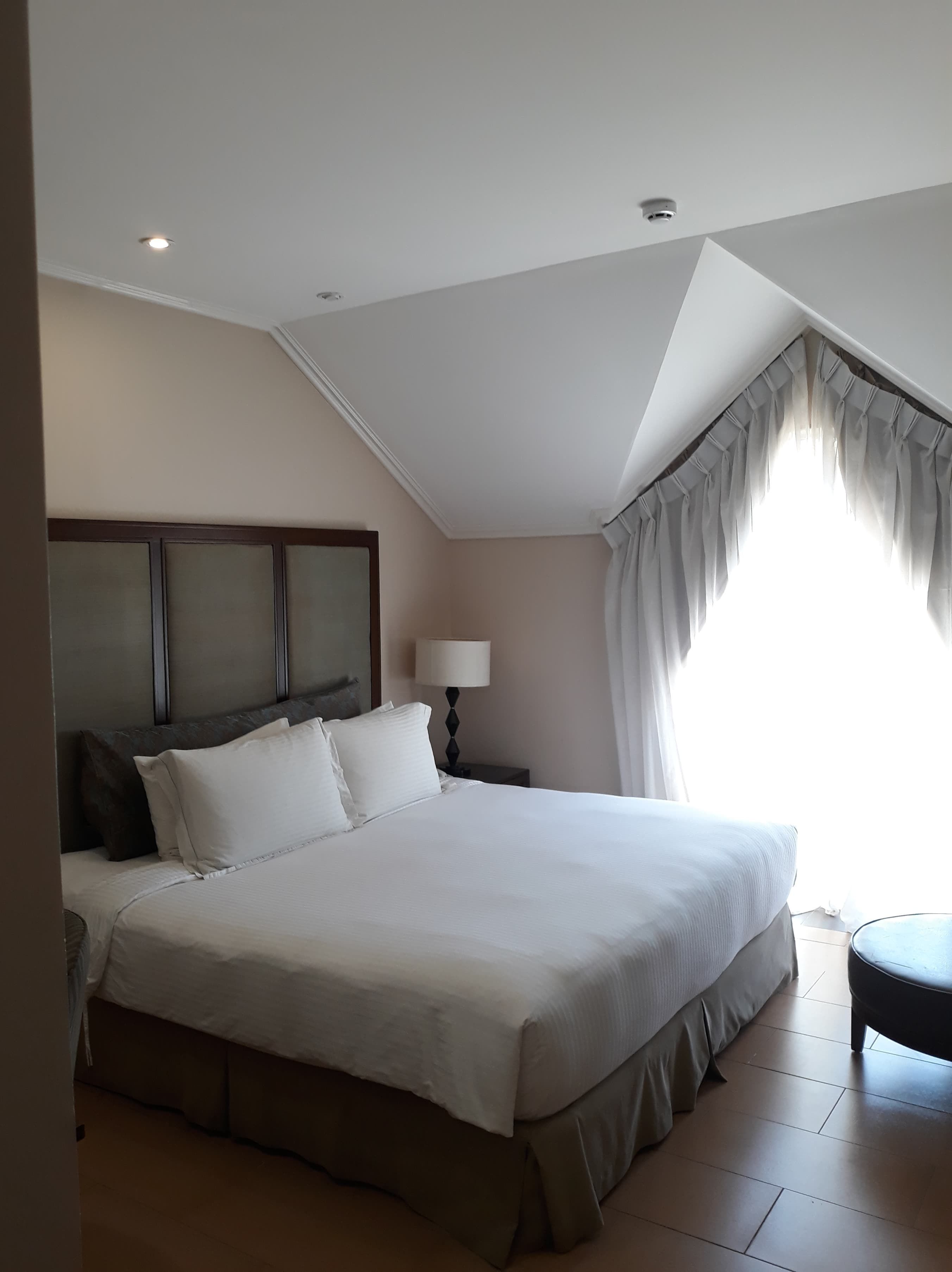 Un dormitorio con una cama grande, una mesita de noche con lámpara, una ventana con cortinas e iluminación en el techo.