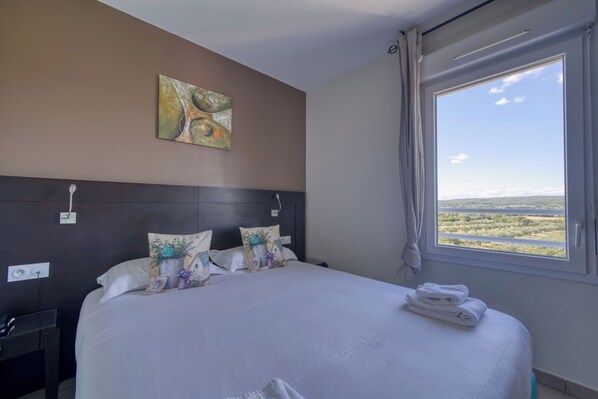 A hotel room with a large bed, a headboard, a painting, a window with a view, and a nightstand.