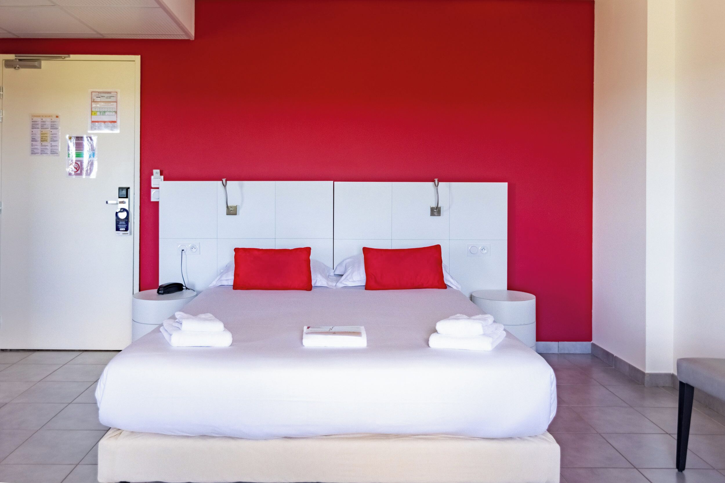 A hotel room with a bed, two bedside tables, a red wall, and a white door.