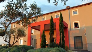 Un edificio moderno con fachada roja, múltiples ventanales y un jardín cuidado con arbustos y árboles.