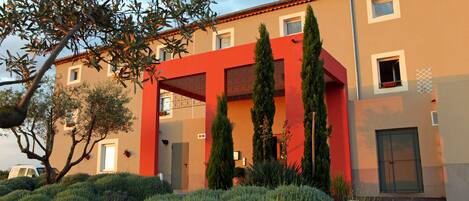 A modern building with a red facade, multiple windows, and a well-maintained garden with shrubs and trees.