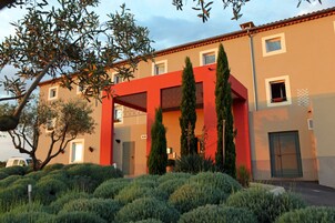 Um edifício moderno com fachada vermelha, várias janelas e um jardim bem cuidado com arbustos e árvores.