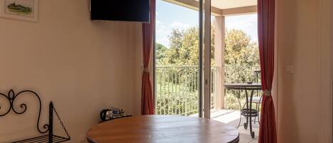 A room with a round wooden table, a TV mounted on the wall, and a balcony with a railing.