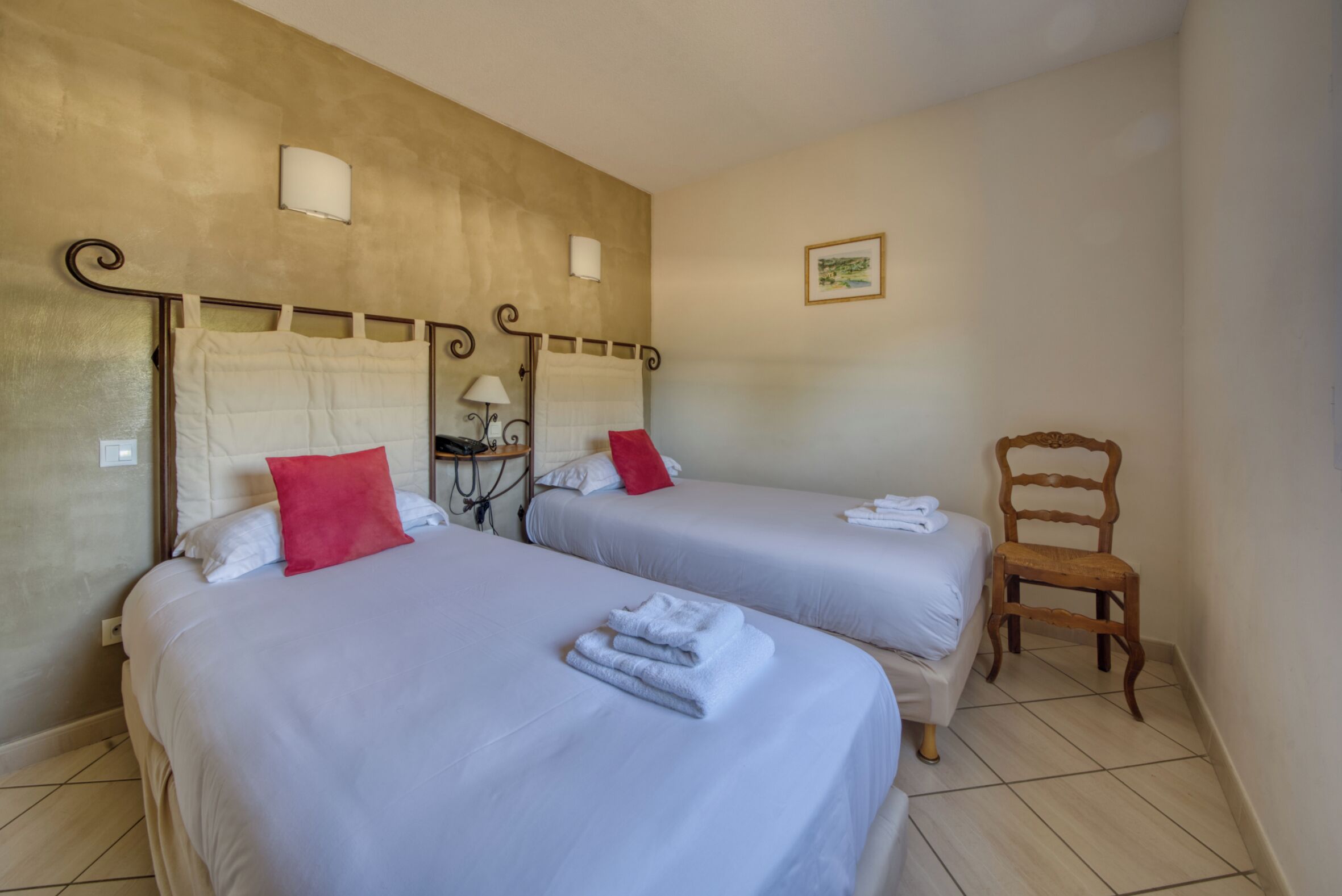 A hotel room with two beds, a chair, and a wall-mounted lamp.