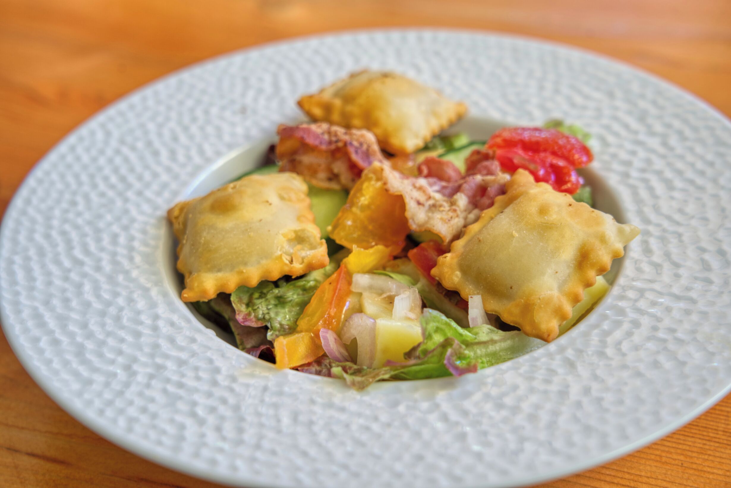 A plate of ravioli with vegetables and bacon on a textured white plate.