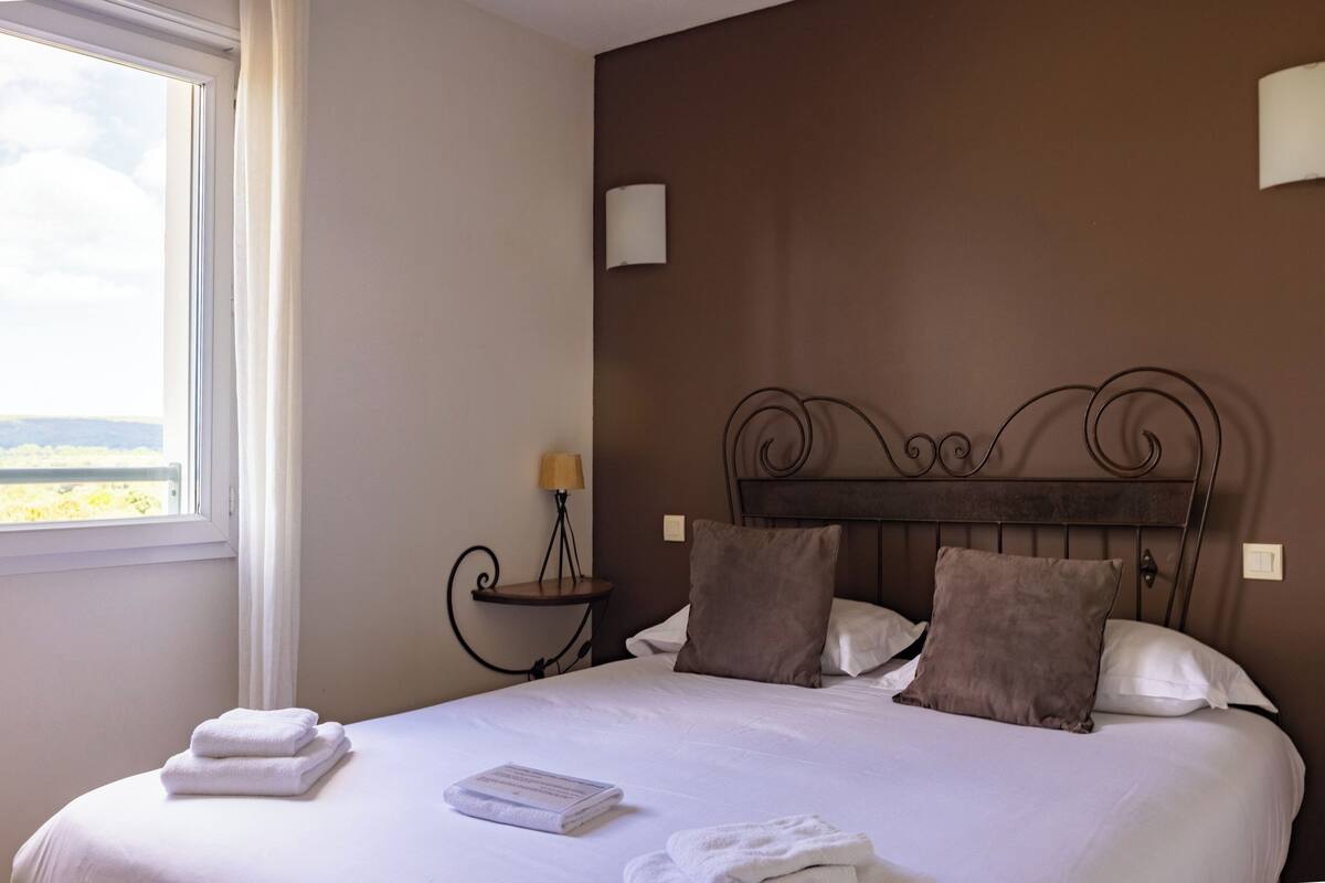 Een net opgemaakt bed met kussens en opgevouwen handdoeken, een nachtkastje met een lamp, en uitzicht op de omgeving door een raam.