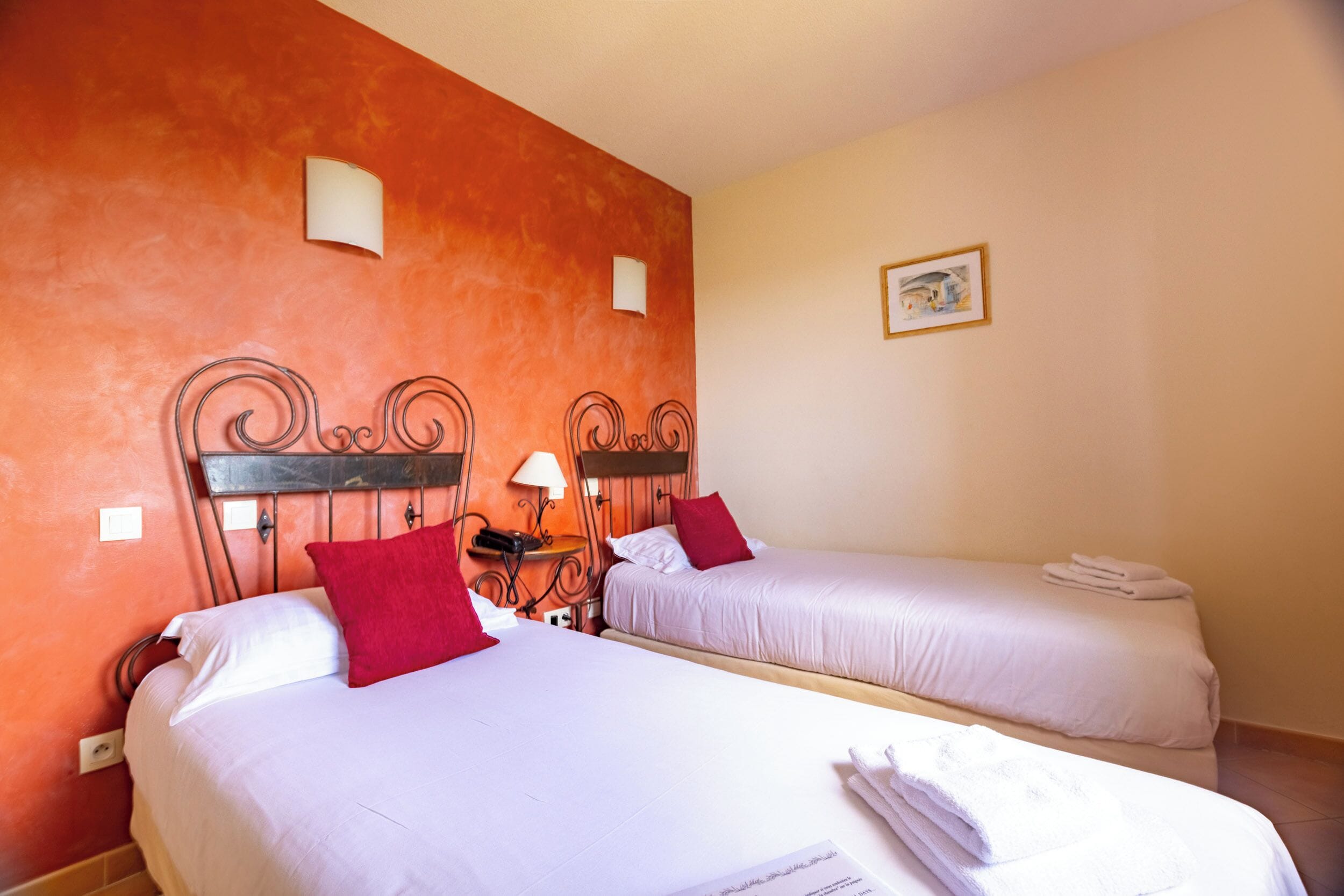 A hotel room with two beds, each with white linens and red pillows, a wooden headboard, a bedside table with a lamp, and a small painting on the wall.