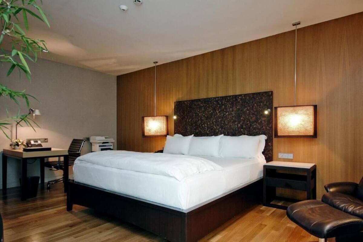 Une chambre d’hôtel moderne avec un plancher en bois, un grand lit, un bureau avec une chaise, un fauteuil en cuir et deux lampes fixées au mur.