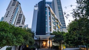 Un hotel moderno con un llamativo letrero 'BUX', rodeado de abundante vegetación y edificios altos contiguos.