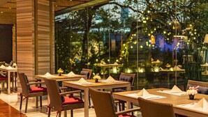 Un comedor interior con mesas y sillas de mimbre, amplias ventanas que ofrecen vistas a árboles y luces de la ciudad.