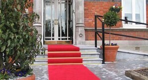 A building entrance with a red carpet leading to a set of stairs, a black metal railing, and potted plants.