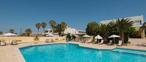 A swimming pool with lounge chairs and umbrellas, surrounded by palm trees and a modern building.
