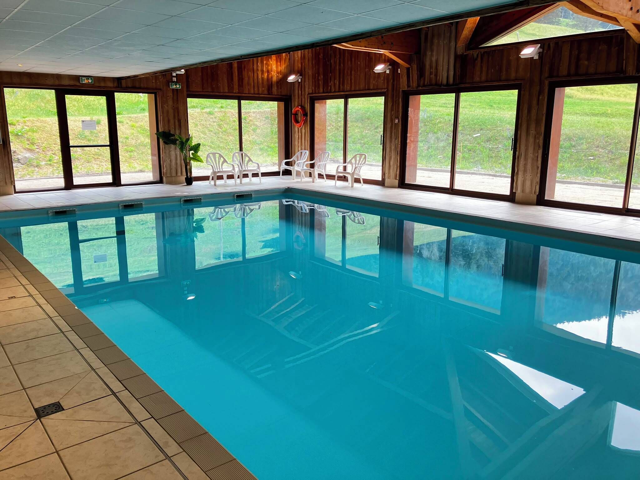 Ein Hallenbad mit großen Glaswänden, Fliesenboden und weißen Kunststoffstühlen.