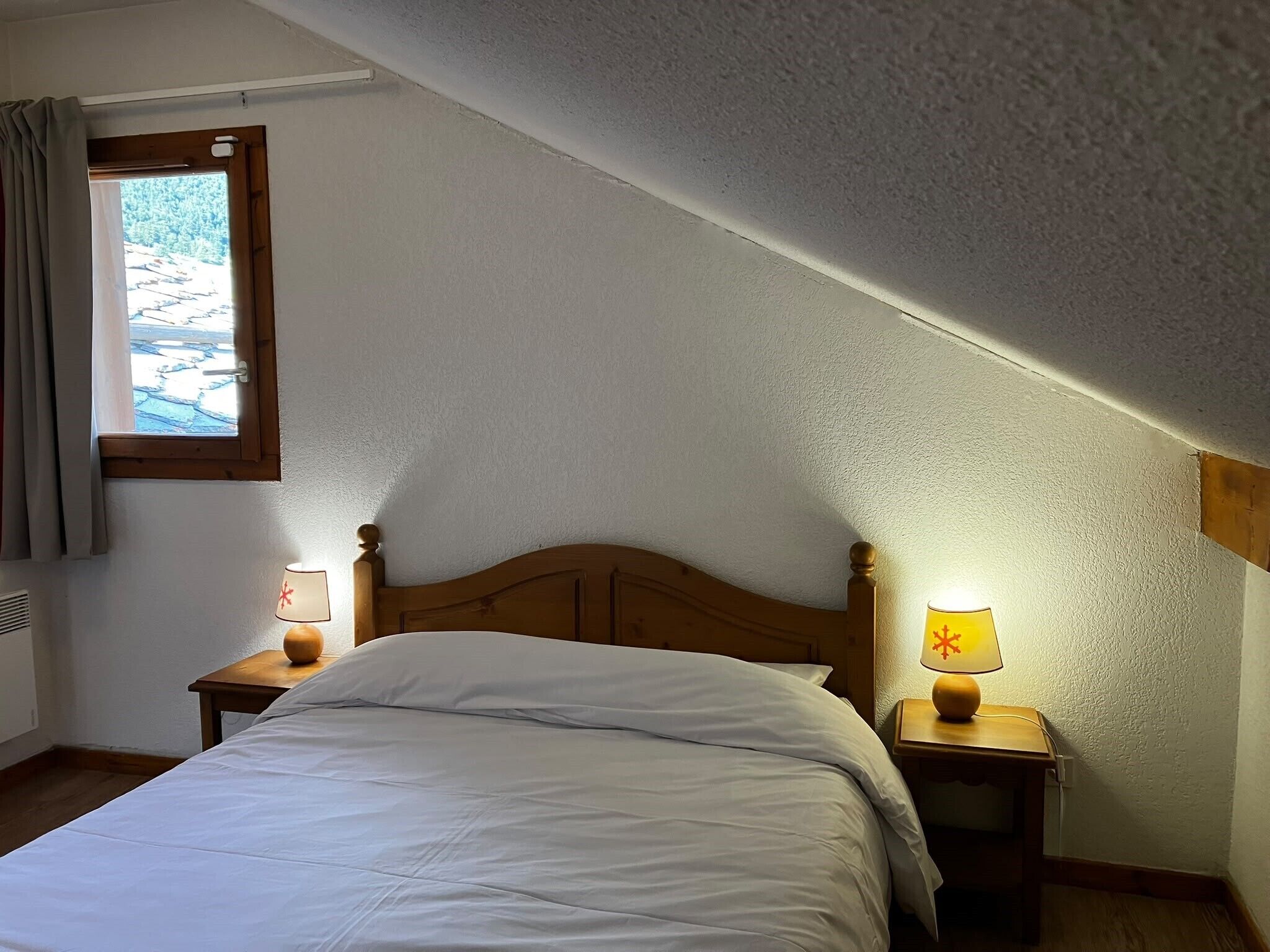 Ein Schlafzimmer mit schrägem Dach, einem Bett, zwei Nachttischen mit Lampen und einem Fenster mit Vorhängen.