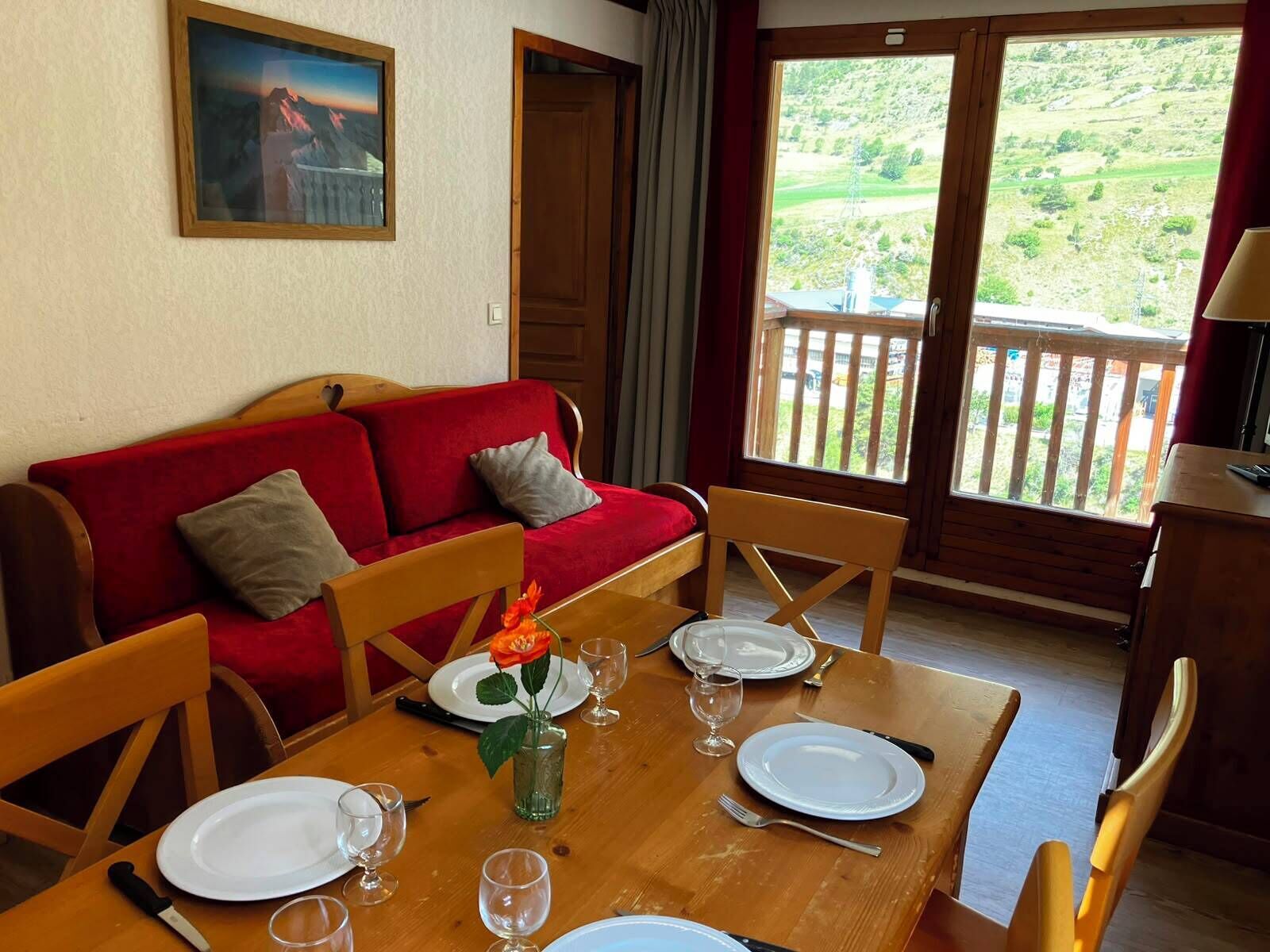 Ein Essbereich mit einem Holztisch, gedeckt für vier Personen, einer roten Couch mit Kissen und Blick auf eine grüne Landschaft durch ein großes Fenster.