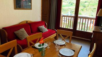 Ein Essbereich mit einem Holztisch, gedeckt für vier Personen, einer roten Couch mit Kissen und Blick auf eine grüne Landschaft durch ein großes Fenster.