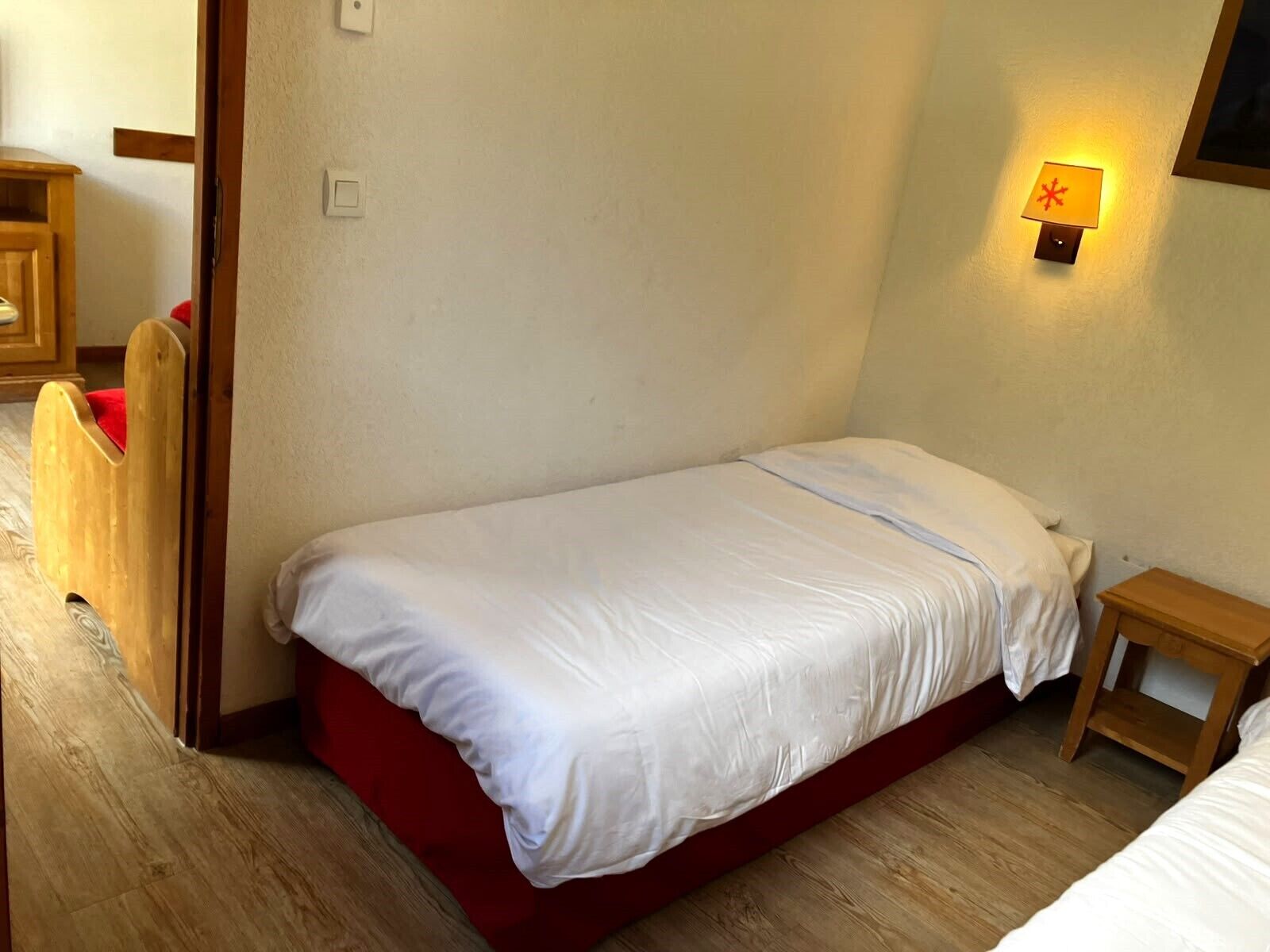 Un lit simple avec une literie blanche, dans une chambre dont le sol est recouvert de parquet et qui est équipée d’une petite table de chevet.