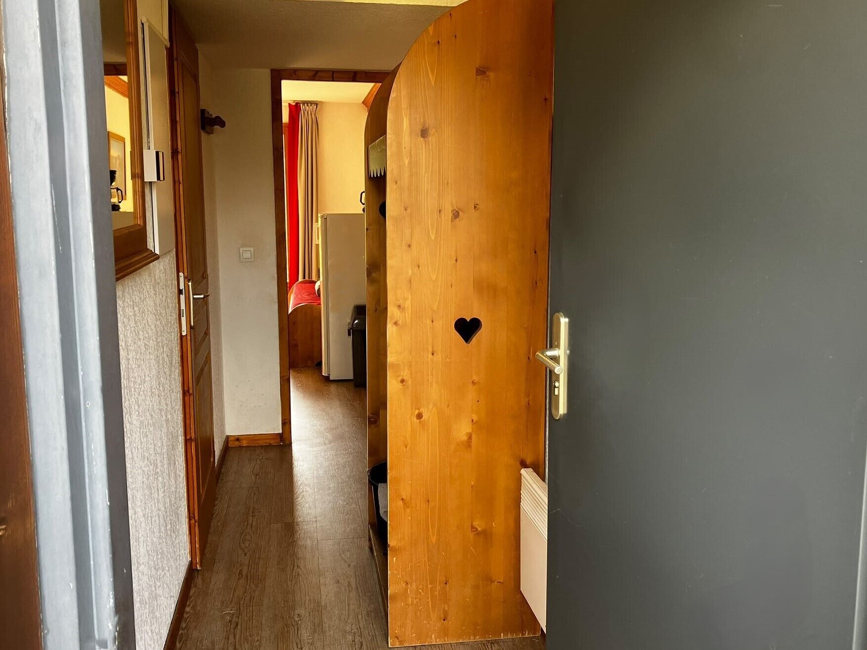 Un pasillo con una puerta de madera que tiene un recorte con forma de corazón, que da a una habitación con radiador.