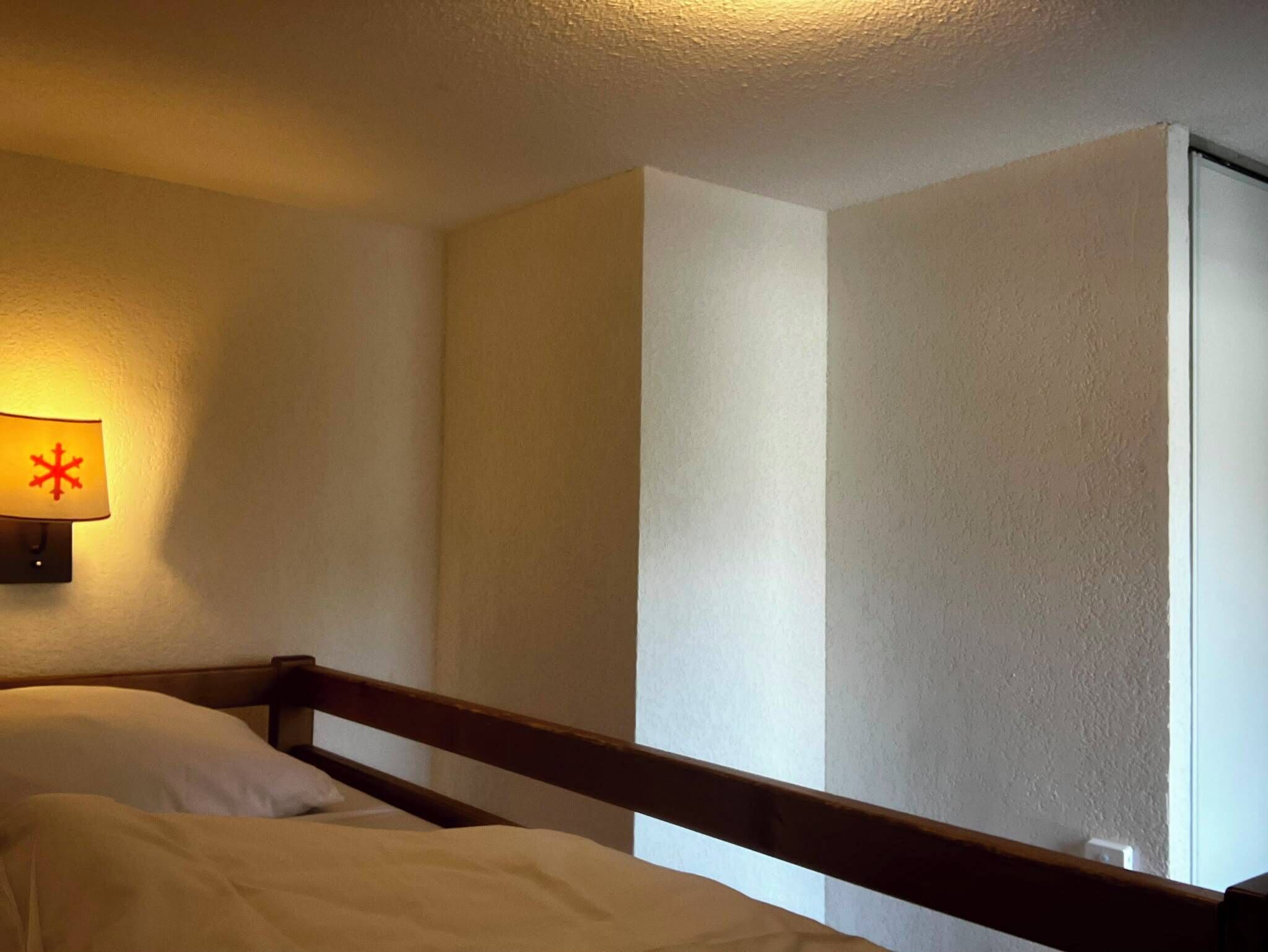 En säng för en person med en trägavel i ett rum med enkla väggar och en enda lampa.