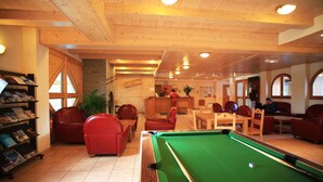 A pool table in a room with wooden ceilings and red chairs. There are bookshelves and a plant in the room.