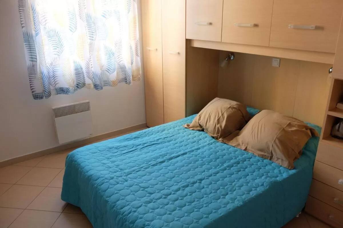 A single bed with a blue quilt, beige pillows, a wooden headboard, and a window with a leaf-patterned curtain.