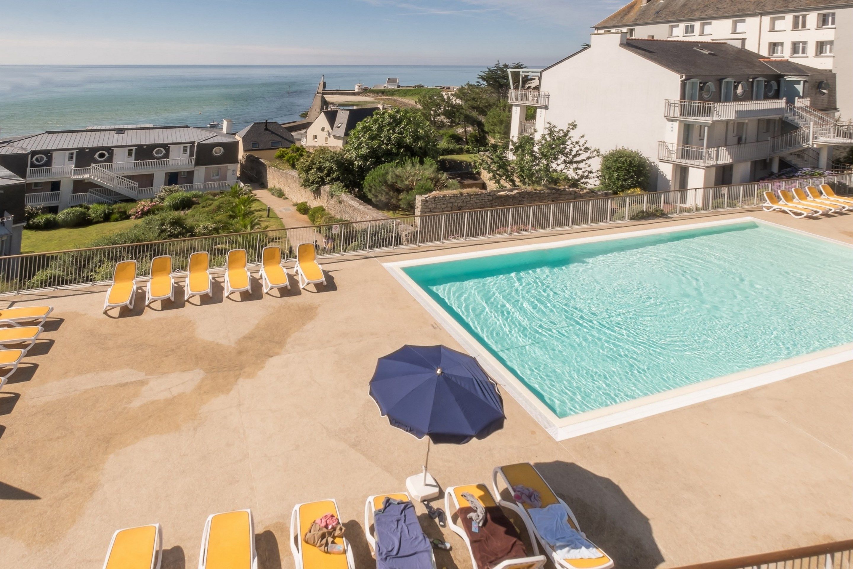 Un espace piscine doté de chaises longues et d’un parasol, offrant une vue sur la côte, avec ses bâtiments et sa végétation.