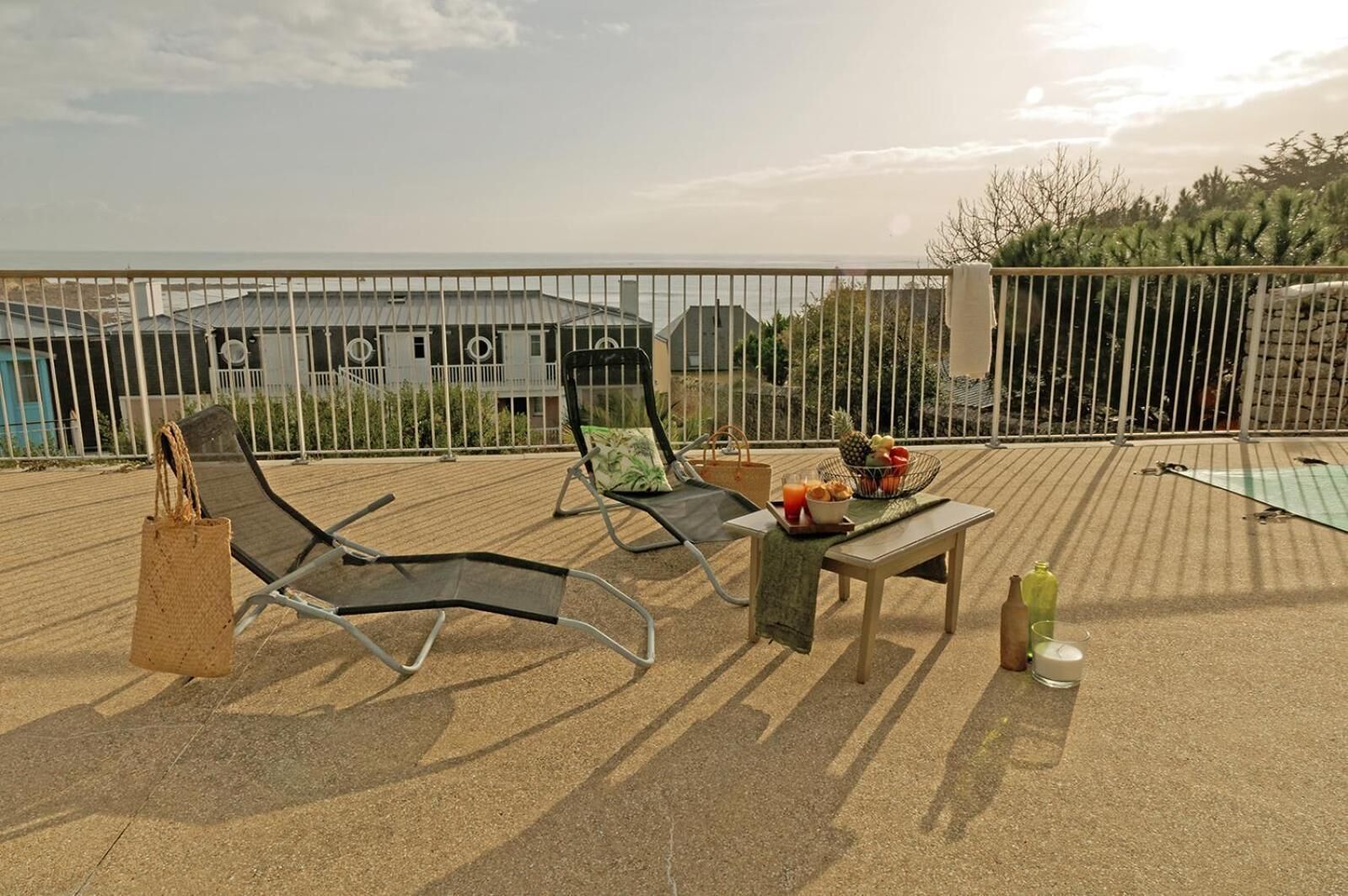 A sunlit outdoor seating area with two lounge chairs, a small table with a drink, and a basket of fruit.