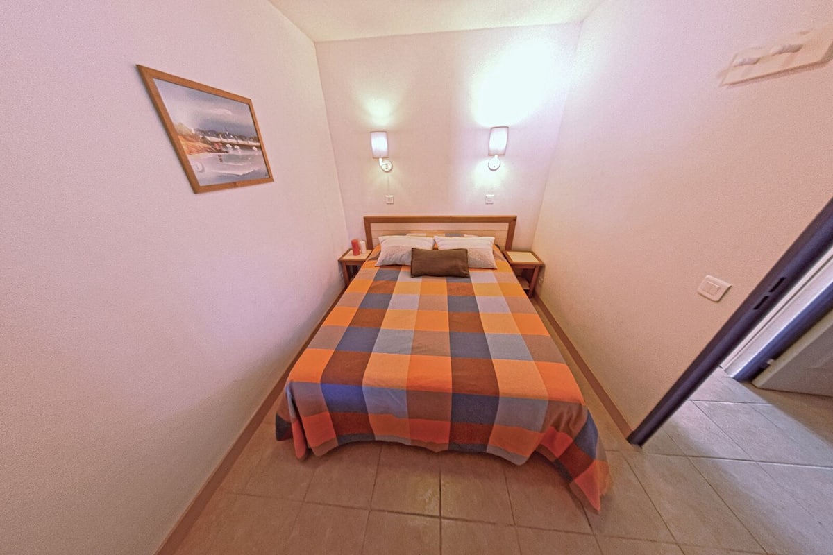 A small, neatly arranged bedroom with a checkered bedspread, two bedside lamps, a nightstand, and a framed picture on the wall.