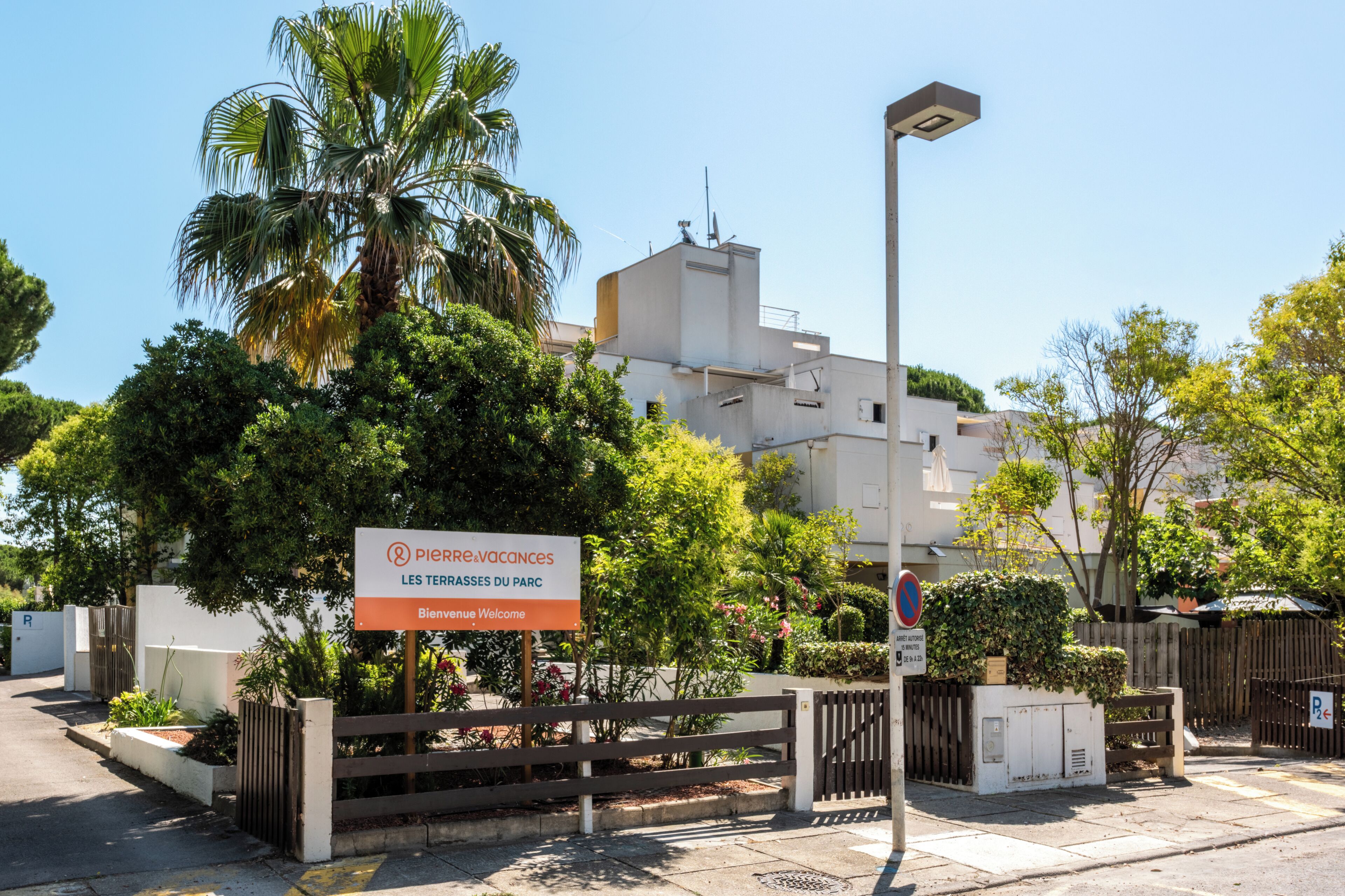 Une enseigne indiquant « Piers Vacances Les Terrasses du Parc », accompagnée d’un message d’accueil, d’une entrée fermée par un portail et d’un lampadaire.