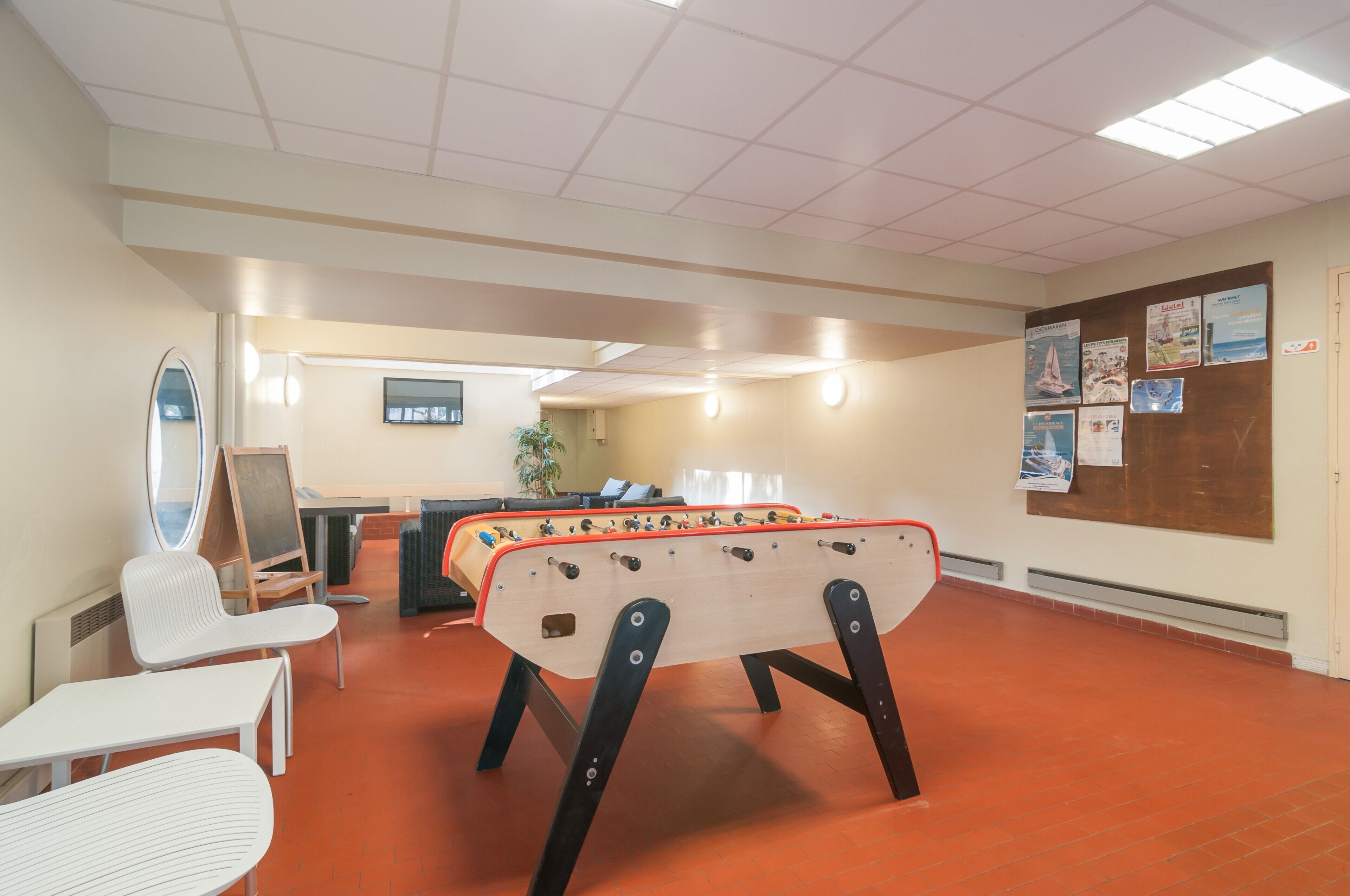 Une pièce avec une table de baby-foot, des chaises blanches, une petite table et un téléviseur.