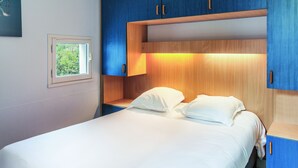 A bedroom with a bed, a nightstand, a window, and built-in wooden cabinets. - Résidence Pierre & Vacances Les Terrasses Du Parc (La Grande-Motte)