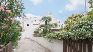 A narrow concrete pathway flanked by wooden fences and lush greenery, leading to a white building with multiple windows and balconies. - Résidence Pierre & Vacances Les Terrasses Du Parc (La Grande-Motte)