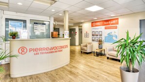 A reception desk with a sign reading 'Pierre Vacances Les Terrasses Du Parc.' There are potted plants, a seating area with chairs, and a wall with framed pictures and a map. - Résidence Pierre & Vacances Les Terrasses Du Parc (La Grande-Motte)