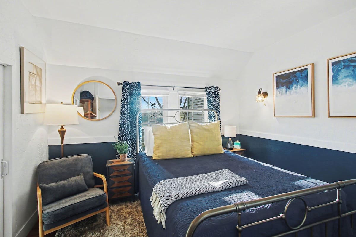 A bedroom with a bed, a chair, a nightstand, a mirror, and wall art.