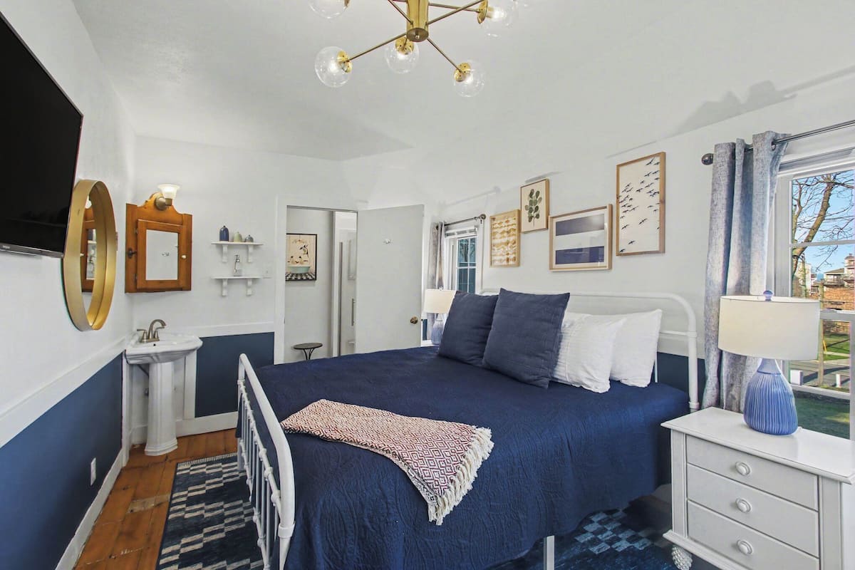 A bedroom with a bed, bedside table, lamp, and a view of the outdoors.